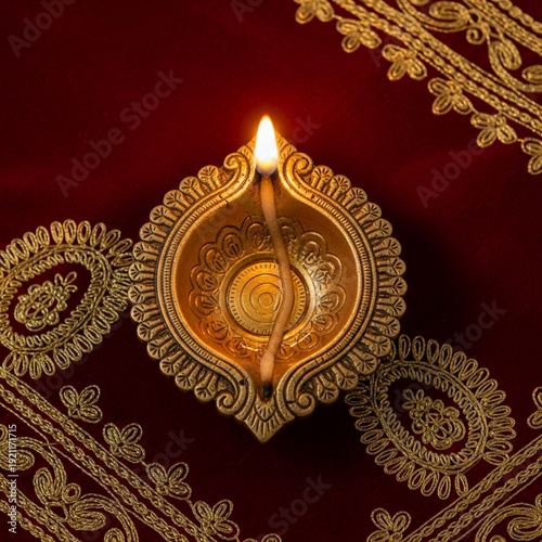 Ornate golden diya lamp with a lit flame on a rich red fabric with intricate gold embroidery, celebrating Diwali and light.