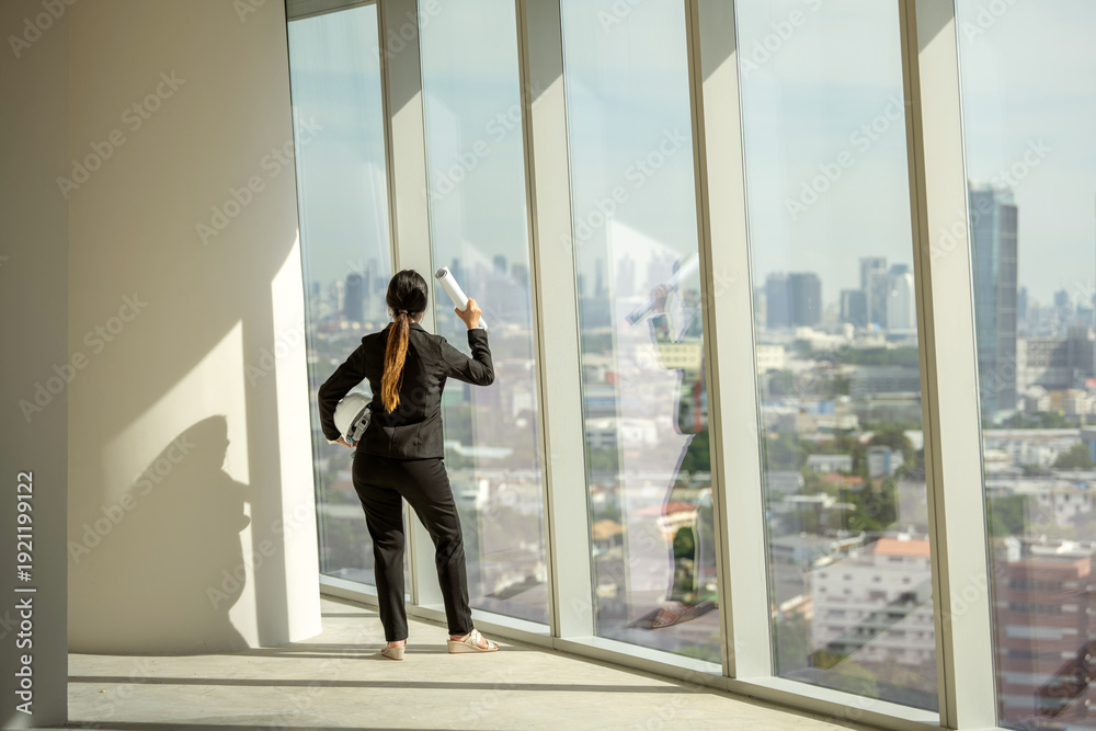 Fototapeta premium architect woman standing raise arms with blueprint helmet victory celebrate success proud empowered achievement on empty office interior near glass window. office architect raise arms empowered
