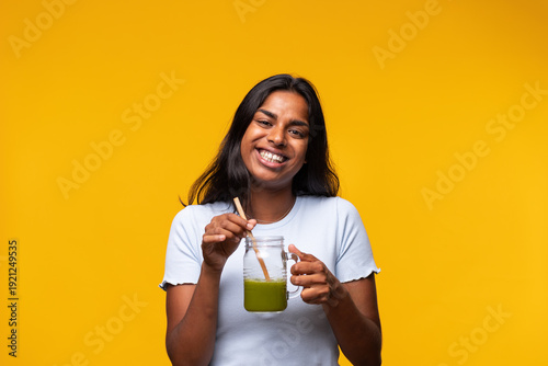 Young woman enjoying green smoothie with reusable straw on vibrant yellow background