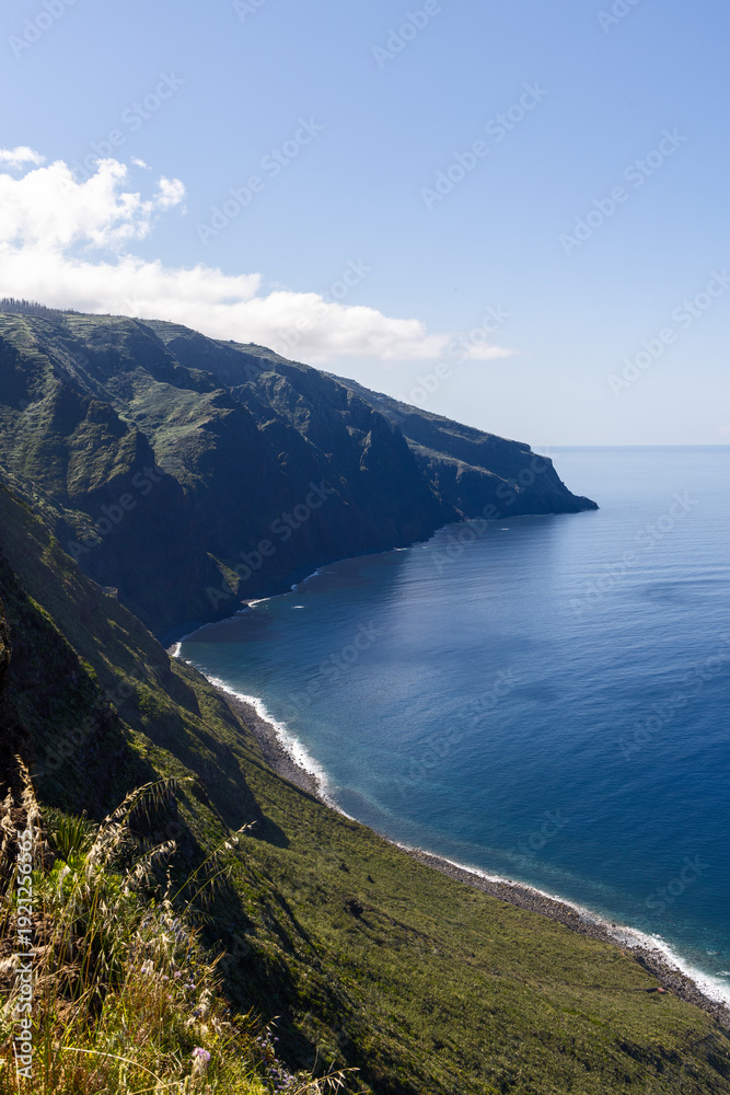 Fototapeta premium Ponta do Pargo Atlantic cliffs Madeira Portugal vertical coastline view, steep green volcanic slopes descend toward calm blue ocean with narrow surf edge and vast clear sky