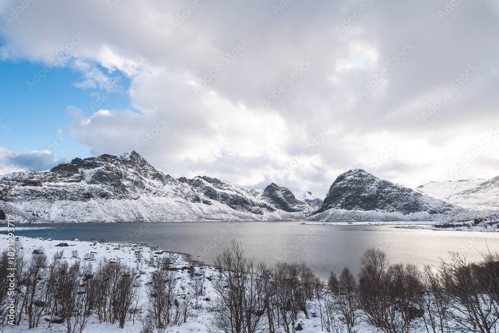 Obraz premium Sunlight breaks through heavy clouds over the snowy fjords near Sund, Lofoten, Norway, illuminating a lone red cabin in a dramatic Arctic winter landscape