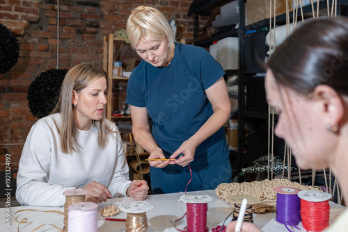 Explaining craft mentor showing proper yarn handling and hook movements while students practice stitches at table indoors.