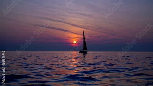 Sailboat moves gracefully over tranquil waters during sunset, with colorful reflections illuminating the surface and enhancing the peaceful scene