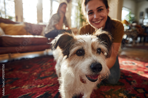 Visiting veterinarian kneels smiling behind a scruffy white - and - brown terrier on a red patterned rug in a sunlit cozy living room during home visit