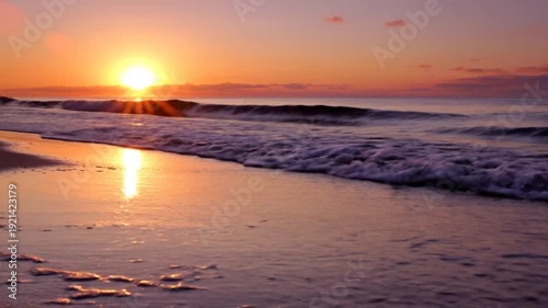 Serene ocean waves gently lap onto a sandy beach at sunset, with the sun casting a warm golden glow across the sky and water.