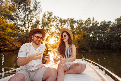 Couple celebrating anniversary opening a bottle of champagne while sailing on a boat in sunset