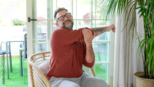 Obraz na plátně Active mature bearded man exercising inside home with stretching exercises for shoulders and arms in a modern living room