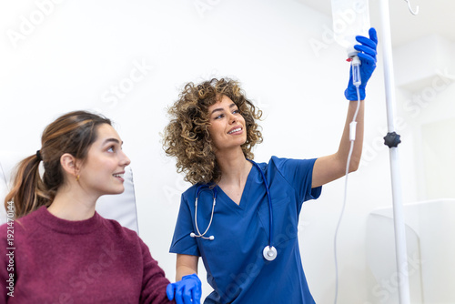 Obraz na plátně A curly-haired doctor in blue scrubs looks up while adjusting a clear IV infusion bag on a stand