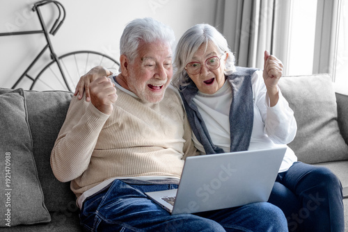 Foto A happy elderly couple, sitting on the sofa in a modern home, embrace while using their laptops, excited by some good news and spending carefree, cheerful, and harmonious moments together
