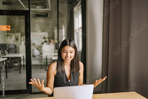 Young mixed race woman expressing excitement while engaging in a virtual laptop meeting in a modern office setting during daytime