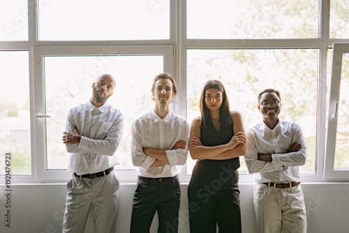 A diverse group of four professionals standing confidently by a large window in a contemporary office, dressed in business attire and displaying collaboration and unity