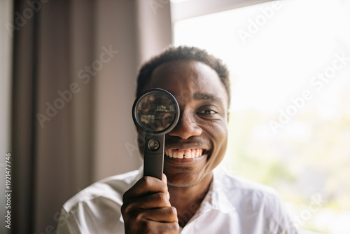A cheerful black man smiles brightly while holding a magnifying glass up to his eye, immersed in curiosity.