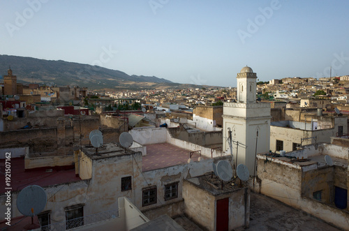Rooftop view of the old medina in Fez, Morocco, with satellite dishes, aged buildings, and distant hills under a clear evening sky