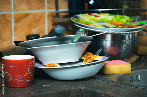 A messy kitchen counter featuring a chaotic stack of dirty white bowls plates and a red mug with food residue sitting next to a yellow cleaning