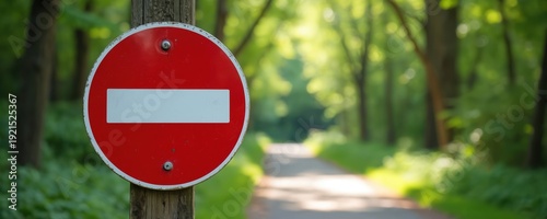 Red no entry sign attached to wooden post on forest path. Road closed for vehicles. No passage allowed through woods. Green trees and bright sunlight.