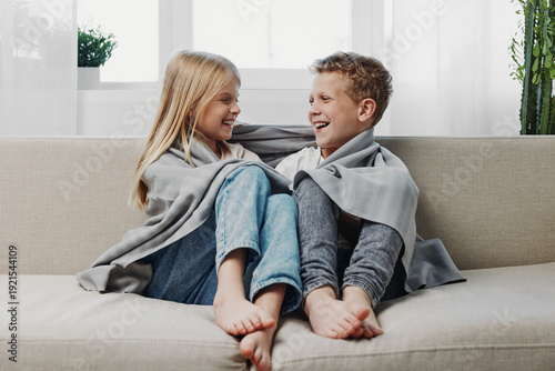 Quadro su tela Happy children wrapped in grey blanket sitting barefoot on couch smiling and looking at each other in bright living room