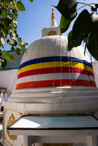 Quadro em tela BARBAR, BAHRAIN - FEBRUARY 16, 2026: A white Buddhist stupa adorned with the colorful striped Buddhist flag and fairy lights, framed by bodhi tree leaves at a local temple