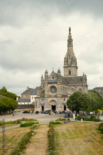 View of the Basilica of Sainte-Anne d'Auray in Brittany, France. The stone neo-Gothic facade features a tall central tower, a large rose window, and ornate sculptures under a dramatic cloudy sky