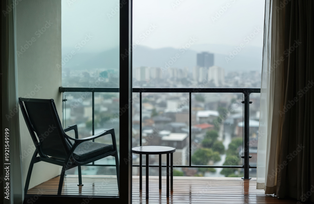Fototapeta premium View from apartment window on rainy city with balcony chair and table. Modern buildings and distant mountains in fog. Overcast sky, grey weather. Urban landscape.