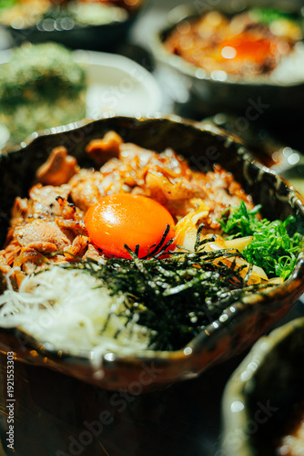 Close-up of a traditional Japanese beef rice bowl (donburi) topped with fresh raw egg yolk, shredded nori seaweed, green onions, and caramelized onions. Served in a rustic ceramic bowl with grilled po
