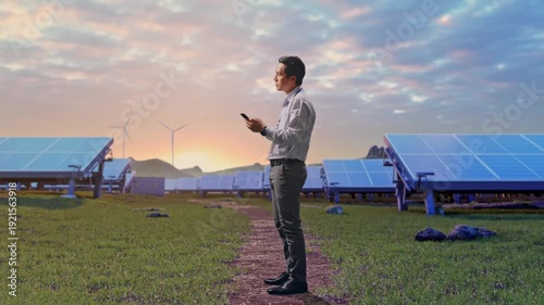 Full Body Side View Of Asian Male Worker Standing With His phone with Solar Panels and Wind Turbines on a Green Field, He Observes By Looking Around Before He Come To Concentrating With His smartphone