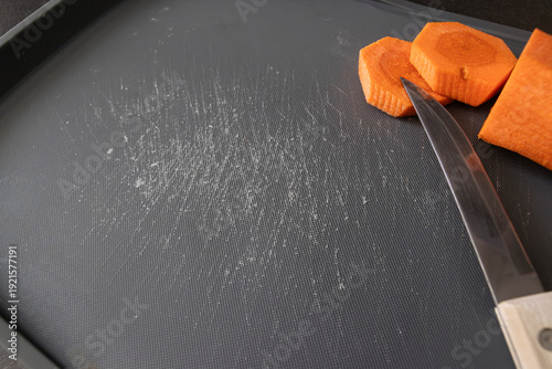 A kitchen knife cuts carrot pieces on a damaged plastic board with deep cut marks. Concept of microplastic contamination, unsafe kitchen tools, environmental impact, material degradation.