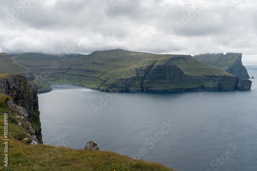 A panoramic view of the secluded village of Tjørnuvík nestled along a quiet bay and surrounded by dramatic mountains viewed from a high-altitude vantage point