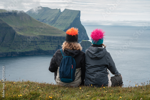 Young adult couple sitting on cliff looking at Mylingur peak and dramatic mountains, Faroe Islands 
