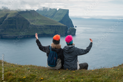 Young adult couple sitting on cliff and enjoying view of Mylingur peak and dramatic mountains, Faroe Islands