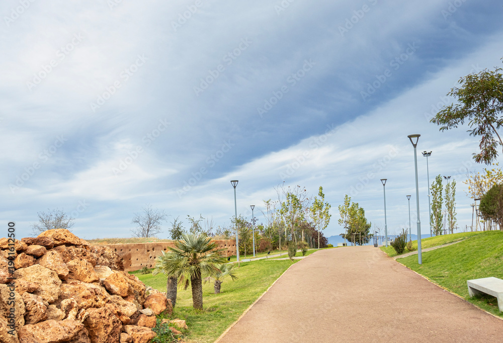 Obraz premium Scenic Modern Park Pathway with Palm Trees and Natural Rock Formations in Morocco under Blue Sky