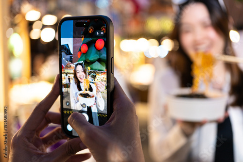 Happy beautiful asian woman posing with street food while a friend takes her photo with a smartphone at a vibrant night market.