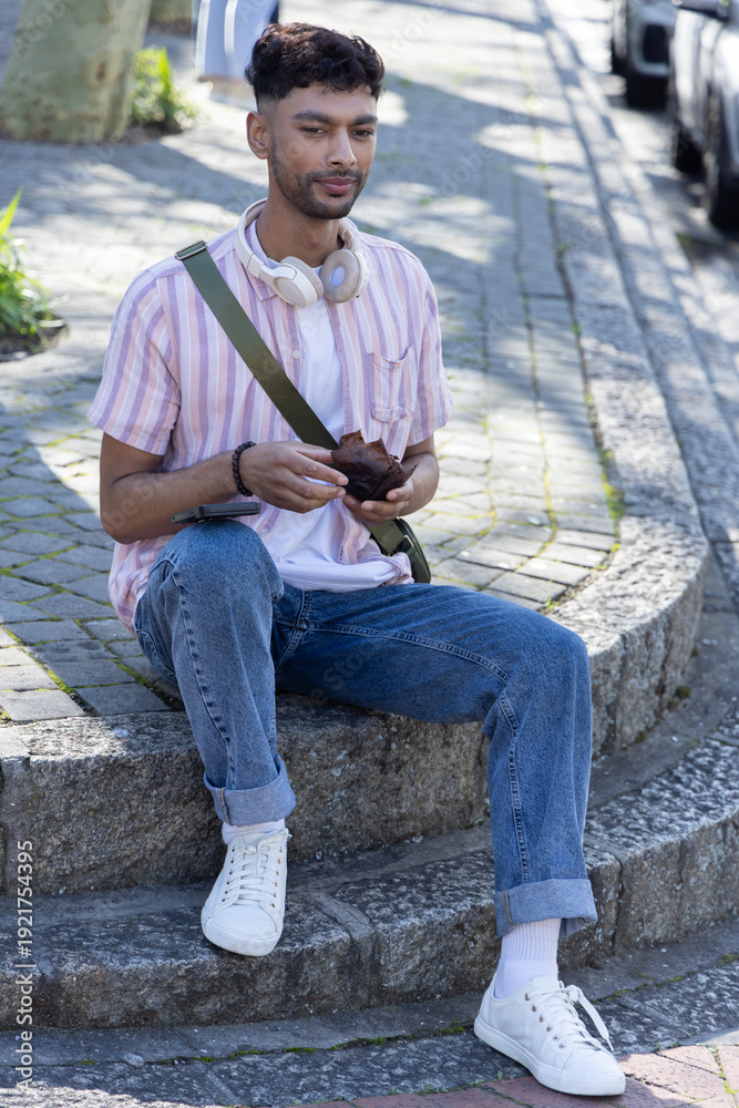Fototapeta premium Asian man sitting on rounded curb holding small brown object while wearing headphones and green bag