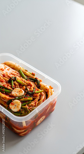 Homemade kimchi packed in a simple transparent plastic container positioned in the lower left of a pale gray countertop, wide empty top and right area as negative space for messaging.
