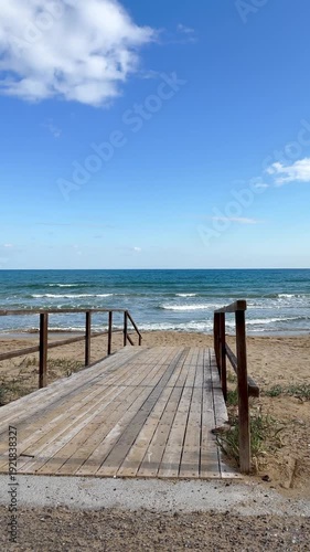 Wooden walkway extending toward the ocean, with waves and a vast blue sea under a bright sky. Minimalist coastal scene highlighting the harmony between natural beauty and human-made structures