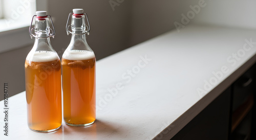 Two plain flip‑top bottles of kombucha standing close together on the left side of a long light stone countertop, large empty right side used as negative space for wellness or beverage branding text.