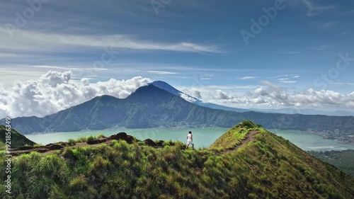 Aerial view sportsman running along narrow green ridge of Mount Batur with panoramic view of Lake Batur and lush volcanic peaks under a clear blue sky. Active sport and recreation. Slow motion shot