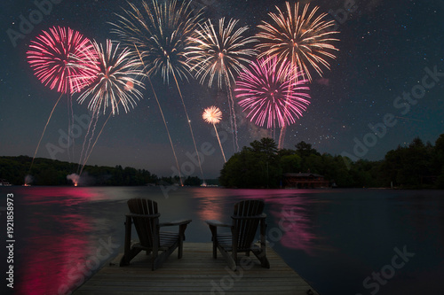 Canvas Print Colorful fireworks exploding in the night sky over a calm lake, with two chairs silhouetted on a dock in the foreground