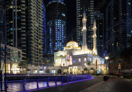 Local sacred skyline beacon illuminated mosque with twin minarets beside Dubai Marina canal and high rise towers at night for urban destination branding and regional identity promotion.