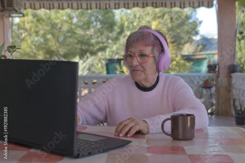 Older woman with headphones happily using her laptop in her country house