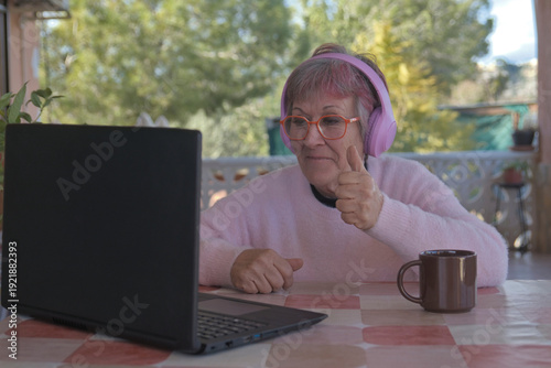 Older woman with headphones happily waving while looking at her laptop in her country house