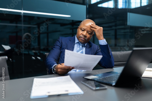 Canvastavla Mature black businessman reviewing printed document while touching forehead in modern office