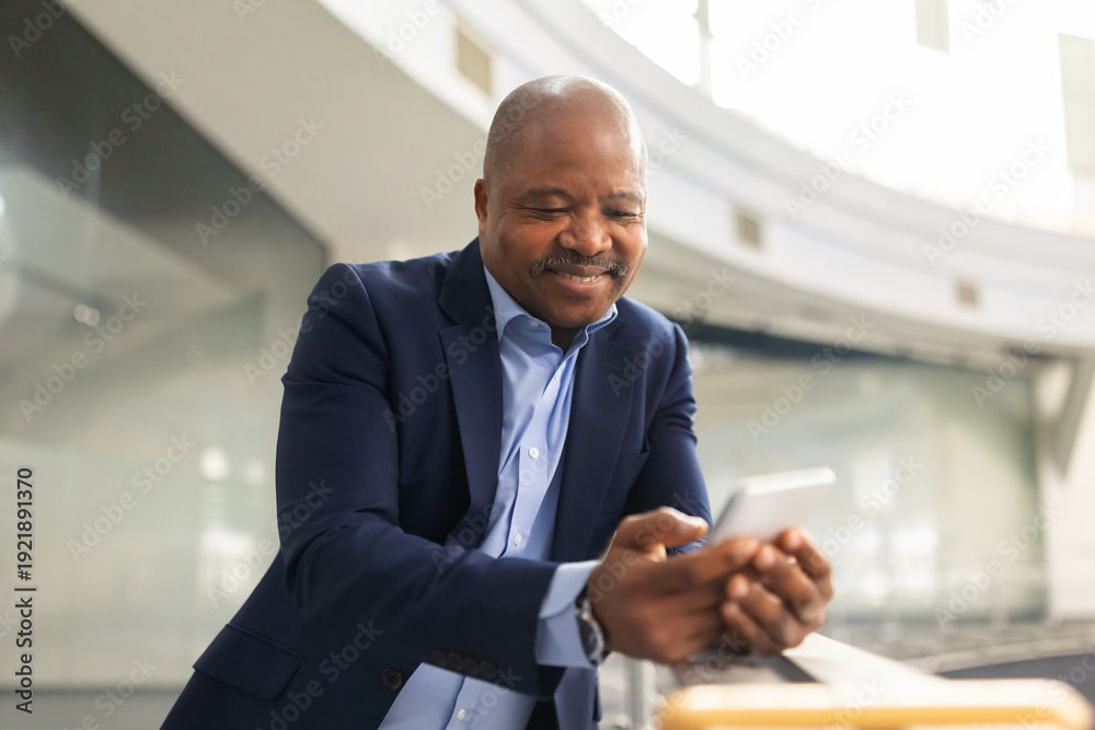 Fototapeta premium Thoughtful mature black businessman looking at smartphone in office atrium, confident corporate leader reviewing information in modern architectural setting