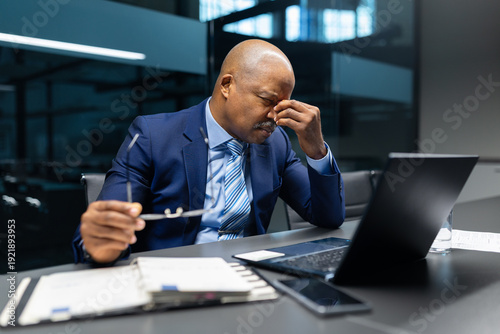 Stressed mature black businessman rubbing eyes while holding glasses and looking at laptop in office. Executive experiencing work pressure and fatigue