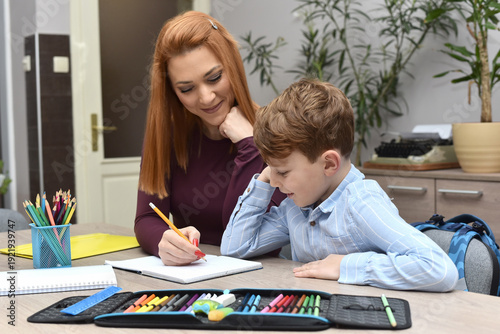 Mother helping her young son with homework at home. Woman and child sitting at a desk, writing in a notebook together. Concept of education, parenting, learning, and family support.