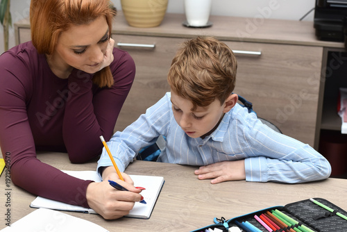 Mother helping her young son with homework at home. Woman and child sitting at a desk, writing in a notebook together. Concept of education, parenting, learning, and family support.