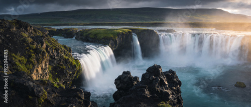 Panoramic view of Godafoss waterfall in Iceland with powerful cascading water, misty spray, dark volcanic rock formations, lush green moss, and dramatic golden sunset light over the horizon. © YOUSSEF