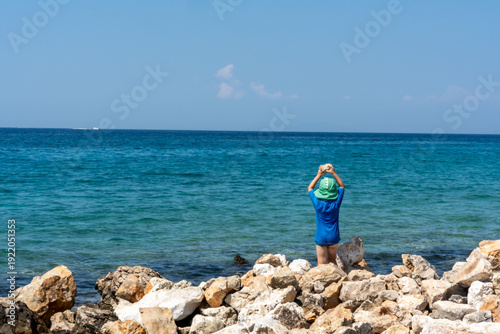 Little boy in blue shirt and green hat throwing stones into turquoise Adriatic sea standing on rocky wild coastline on a bright sunny summer day. Concept of carefree childhood and nature exploration.