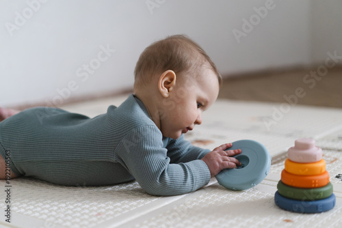 Adorable baby lying on tummy on a soft play mat and playing with a sensory toy. The infant is focused and curious while exploring the toy, supporting early learning and motor skill development.