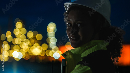 Foto A close up of a female industrial worker in a hard hat and safety gear looking at a lit up factory or refinery at night