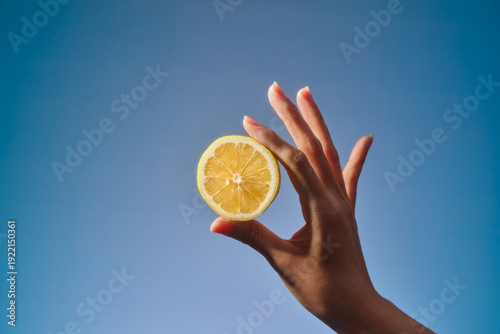 A hand delicately holds a lemon slice against a clear, bright blue sky